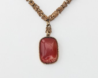Könnte beinhalten: Eine Vintage-Halskette mit einem rechteckigen Anhänger aus rotbraunem Stein. Der Anhänger ist in einen goldfarbenen Metallrahmen gefasst und hängt an einer passenden Kette. Kette und Anhänger haben ein verziertes Design.