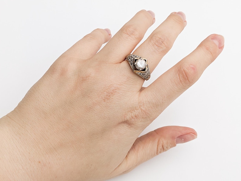 Op de afbeelding: Een gouden ring met een grote, heldere edelsteen gezet in een geometrisch ontwerp. De ring is bezet met kleinere edelstenen rond de hoofdsteen.