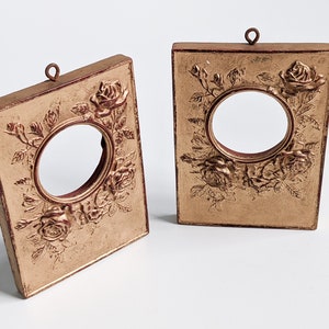 May include: Two gold-colored picture frames with a raised rose design and a circular opening in the center. The frames have a red-brown border and are hanging from a small loop at the top.