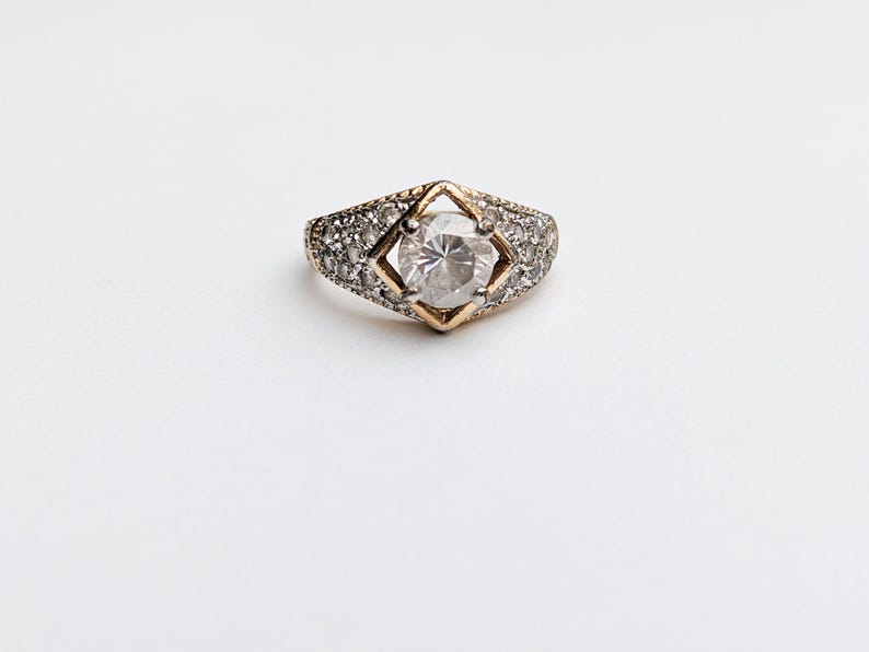 Op de afbeelding: Een gouden ring met een grote, heldere steen in het midden en kleinere, heldere stenen rond de band.