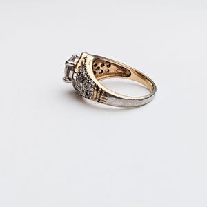 Op de afbeelding: Een gouden en zilveren ring met een grote, heldere steen in het midden en kleinere stenen rond de band.