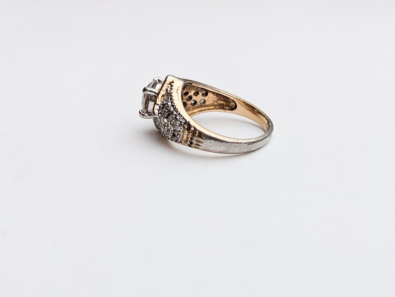 Op de afbeelding: Een gouden en zilveren ring met een grote, heldere steen in het midden en kleinere stenen rond de band.