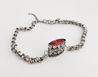Könnte beinhalten: Eine silberfarbene Choker-Halskette mit einem zentralen Anhänger. Der Anhänger hat einen großen, ovalen, roten Edelstein, der von kleineren, klaren Steinen und dekorativen Metallarbeiten umgeben ist. Die Halskette hat ein kettenartiges Design.