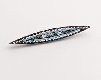 Könnte beinhalten: Eine Vintage-Brosche mit Mosaikdesign. Die längliche, bootsförmige Nadel hat einen schwarzen Rand mit einem sich wiederholenden Dreieckmuster. Die Mitte hat einen hellblauen Hintergrund mit floralen und geometrischen Mosaikelementen.