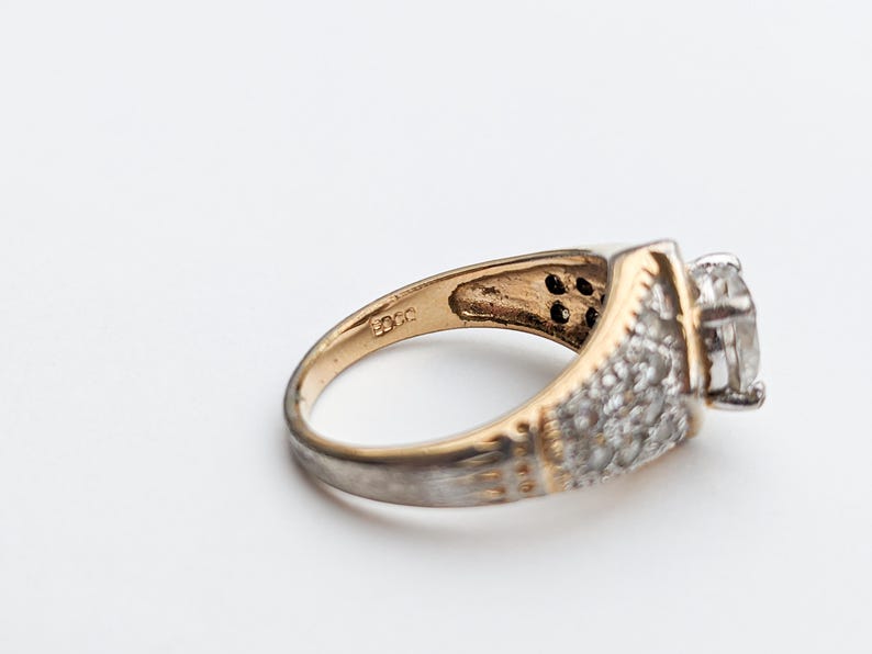 Op de afbeelding: Een gouden en zilveren ring met een grote, heldere steen en kleinere stenen die in de band zijn gezet. De ring is aan de binnenkant gemarkeerd met "925".