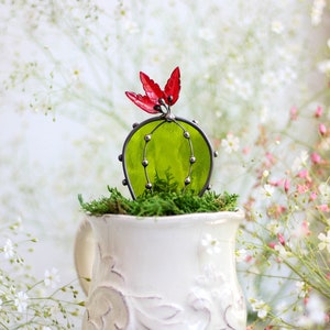 May include: A green glass cactus with red glass flowers sits in a white ceramic mug with a decorative design. The mug is on a silver tray with a white floral background.