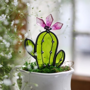 May include: A stained glass cactus with a pink flower on top, sitting in a white ceramic pot with green moss.