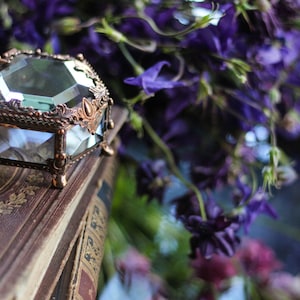 May include: A vintage-style glass and copper-coloured metal jewellery box with a faceted design sits on a stack of old books. The box features a hinged lid and decorative detailing. Purple flowers in the background add colour and softness.
