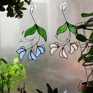 May include: Two stained glass window hangings, one with blue flowers and green leaves, the other with pink flowers and green leaves. Both hangings are attached to suction cups.