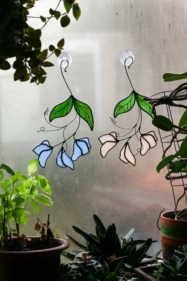 May include: Two stained glass window hangings, one with blue flowers and green leaves, the other with pink flowers and green leaves. Both hangings are attached to suction cups.