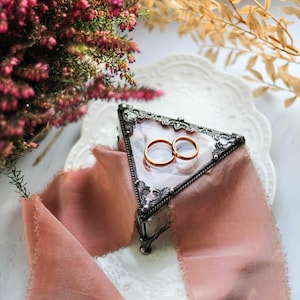 May include: Two gold wedding bands in a triangular glass box with an ornate silver frame. The box is sitting on a white plate with a pink ribbon.