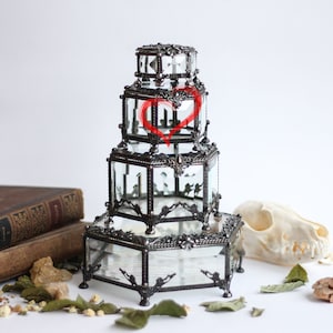 May include: A multi-tiered, vintage-style glass and metal display case with a decorative heart design. The case has a dark metal frame and clear glass panels. The case is on a white surface with a skull, old books, and dried leaves.