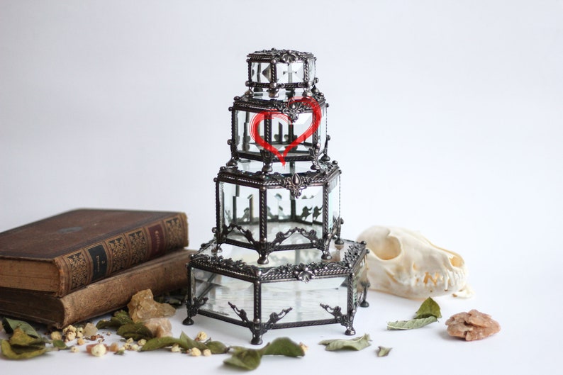 May include: A multi-tiered, vintage-style glass and metal display case with a decorative heart design. The case has a dark metal frame and clear glass panels. The case is on a white surface with a skull, old books, and dried leaves.
