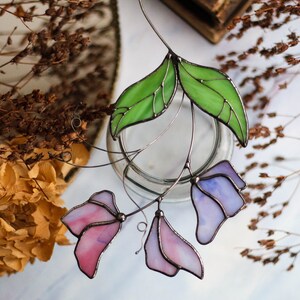 May include: A stained glass floral suncatcher with pink and purple flowers and green leaves. The suncatcher is hanging from a wire.