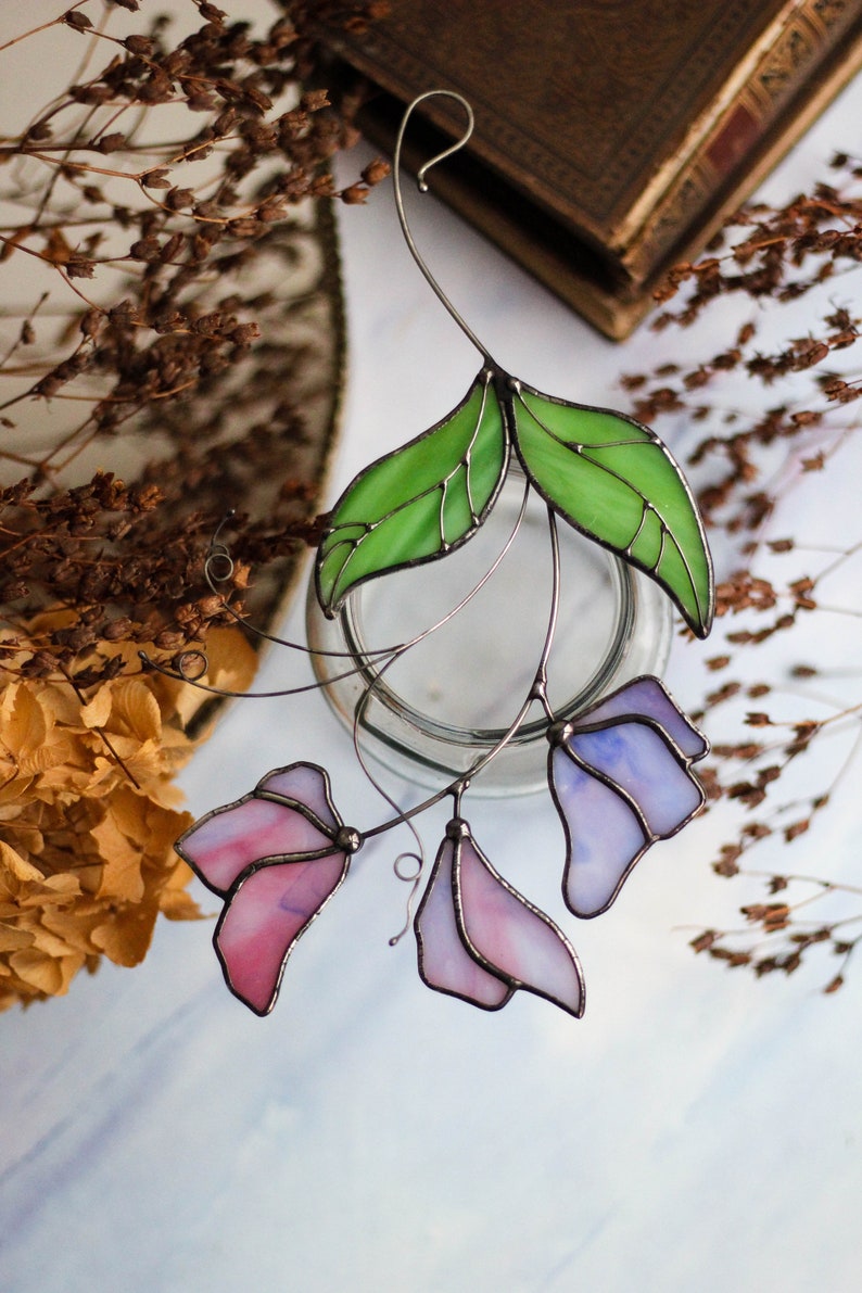 May include: A stained glass floral suncatcher with pink and purple flowers and green leaves. The suncatcher is hanging from a wire.