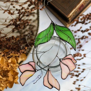 May include: A stained glass floral suncatcher with pink flowers and green leaves. The suncatcher is hanging from a wire and is in front of a glass jar.