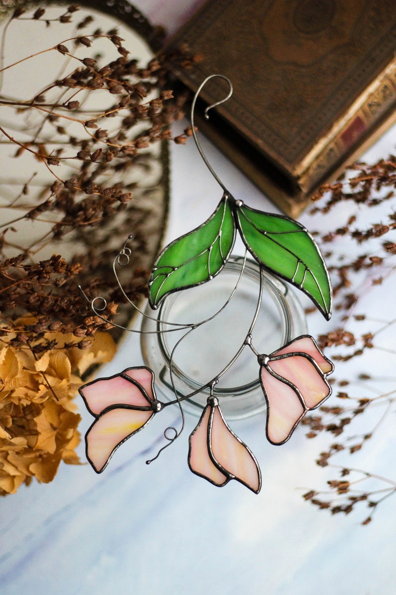 May include: A stained glass floral suncatcher with pink flowers and green leaves. The suncatcher is hanging from a wire and is in front of a glass jar.