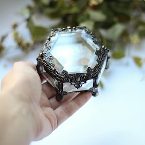 May include: A vintage-style jewellery box made of clear glass and metal. The box has a faceted, hexagonal shape with decorative, dark metal filigree trim. The box is held in a hand, with a blurred green background.