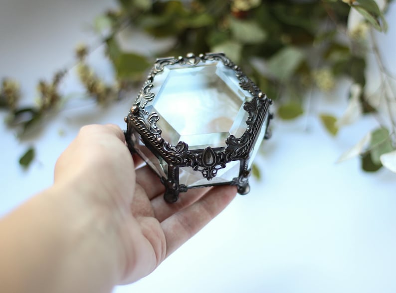 May include: A vintage-style jewellery box made of clear glass and metal. The box has a faceted, hexagonal shape with decorative, dark metal filigree trim. The box is held in a hand, with a blurred green background.