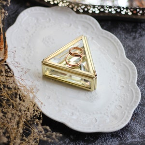 May include: Two gold wedding bands inside a clear glass triangular ring box with a gold trim. The box is sitting on a white plate with a decorative edge.