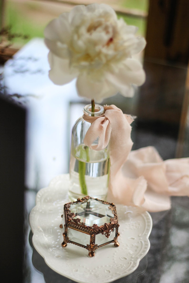 May include: A clear glass bottle with a white peony and a peach ribbon. A small, ornate, hexagonal glass and bronze jewellery box sits on a white, scalloped plate. The scene is soft and romantic.