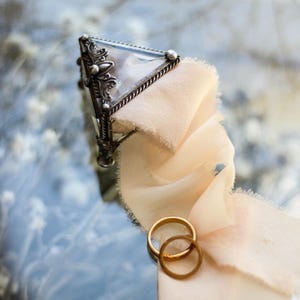 May include: A silver-toned, triangular glass box with ornate detailing, a cream-colored ribbon, and two gold wedding rings. The box is resting on a reflective surface, creating a soft, romantic aesthetic.