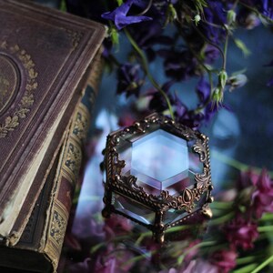 May include: A hexagonal glass and bronze jewellery box with intricate detailing. The box is set amongst purple flowers and an antique book. The box is likely used for storing small items.