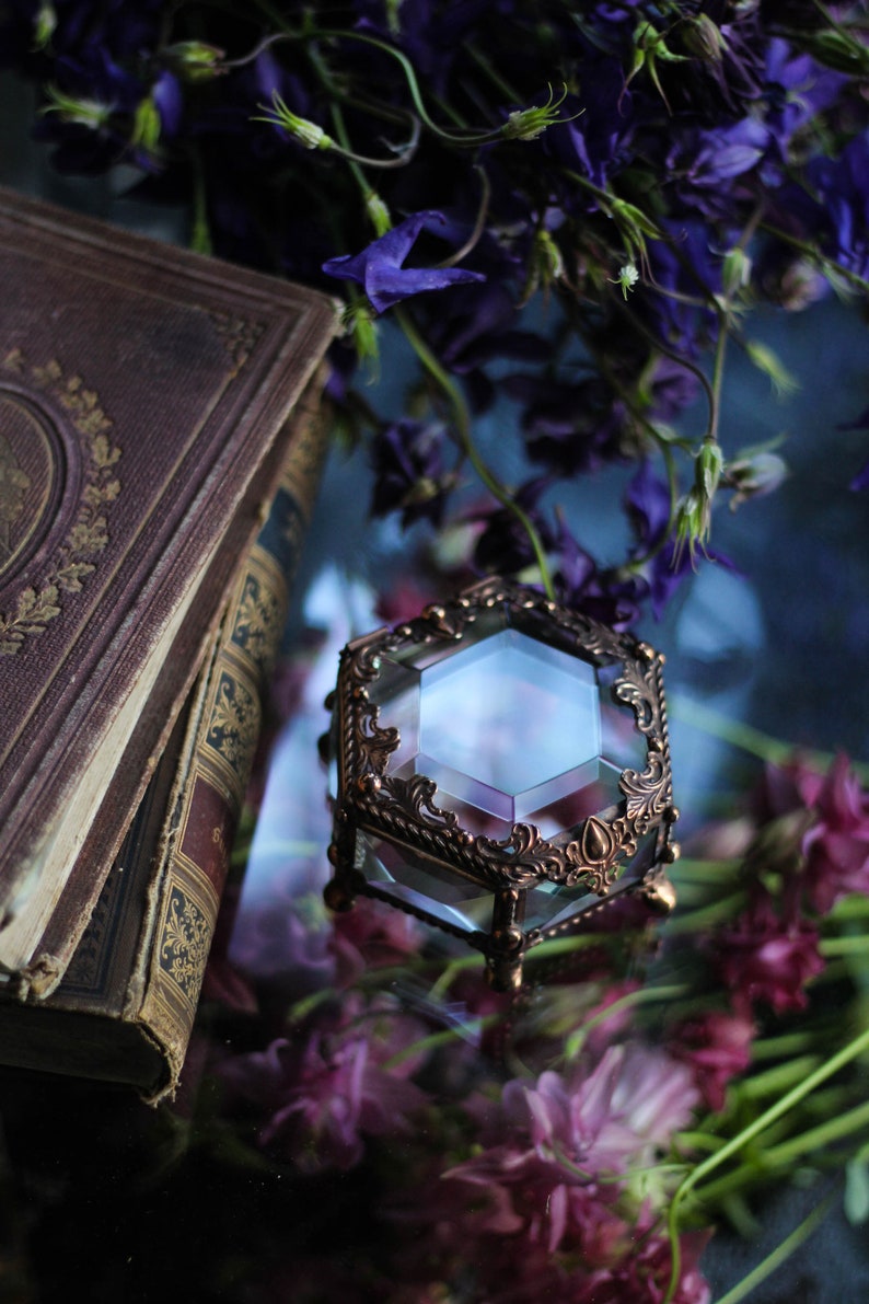 May include: A hexagonal glass and bronze jewellery box with intricate detailing. The box is set amongst purple flowers and an antique book. The box is likely used for storing small items.