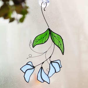 May include: A stained glass window hanging with a suction cup. The design features blue flowers and green leaves.