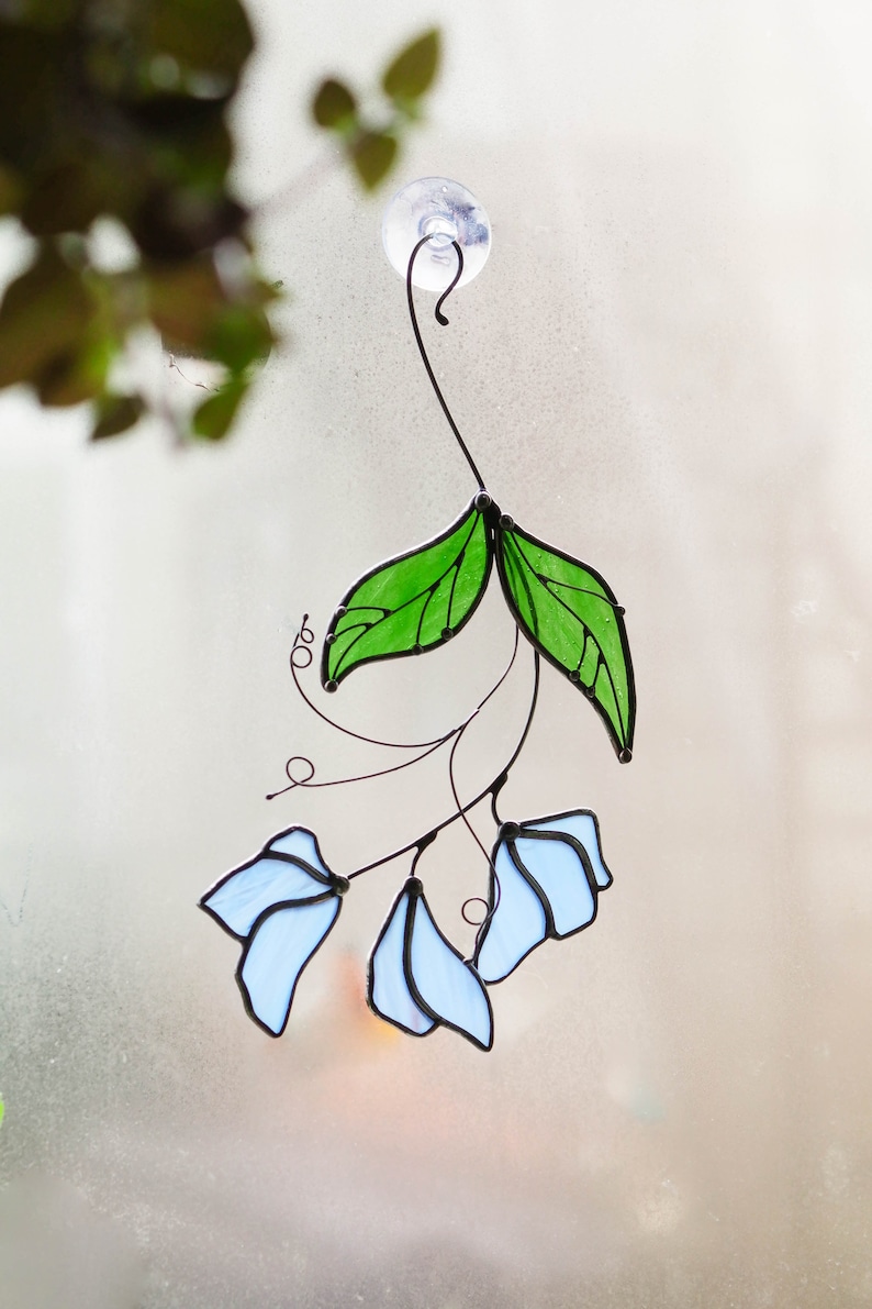 May include: A stained glass window hanging with a suction cup. The design features blue flowers and green leaves.
