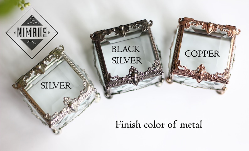 May include: Three small, square glass boxes with ornate metal frames in silver, black silver, and copper finishes. The boxes have hinged lids and are labelled with their respective metal finish colours. The Nimbus logo is in the upper left corner.