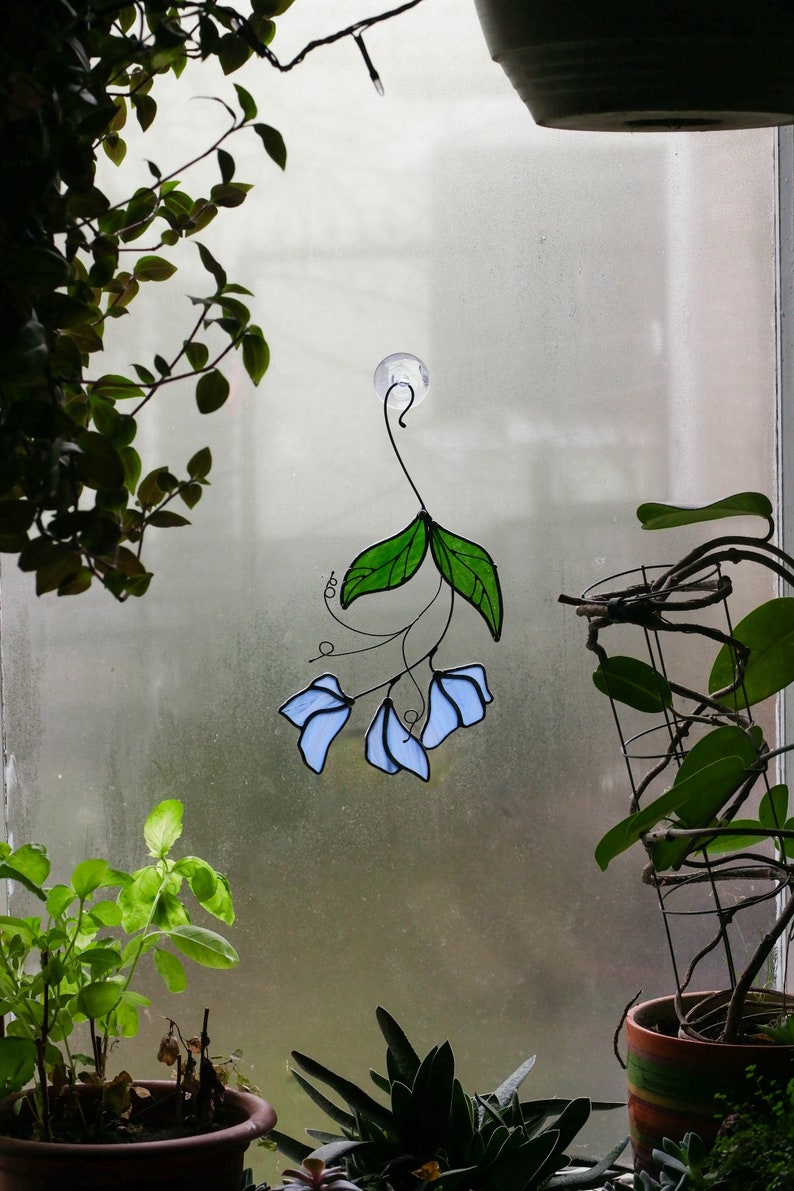 May include: A stained glass window hanging with a blue and green floral design. The hanging is attached to a window with a suction cup.
