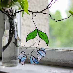 May include: A stained glass window hanging with blue and green flowers. The flowers are made of glass and are attached to a wire frame. The window is hanging in front of a window with a view of green trees.