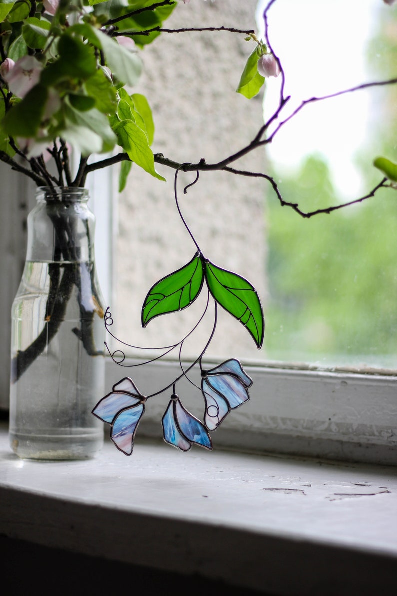 May include: A stained glass window hanging with blue and green flowers. The flowers are made of glass and are attached to a wire frame. The window is hanging in front of a window with a view of green trees.