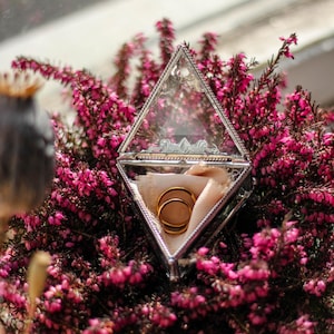 May include: Two gold wedding bands inside a clear glass box with a pink velvet lining. The box is surrounded by pink heather flowers.