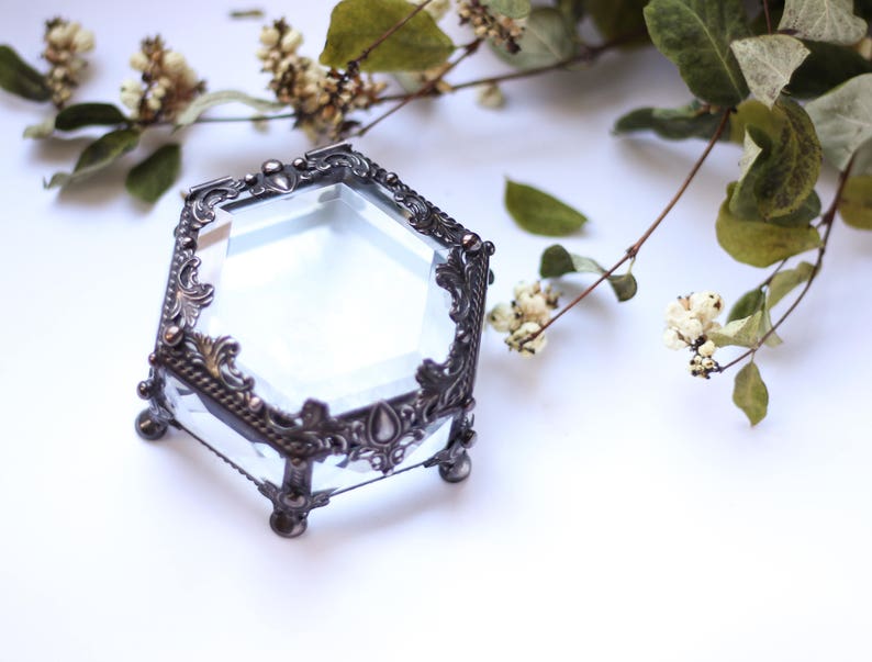 May include: A clear, hexagonal glass jewellery box with ornate, dark metal trim. The box sits on small, rounded feet. Dried flowers and greenery are arranged around the box, creating a vintage aesthetic.