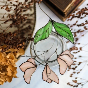 May include: A stained glass floral suncatcher with pink flowers and green leaves. The suncatcher is hanging from a wire and is suspended in front of a white background.