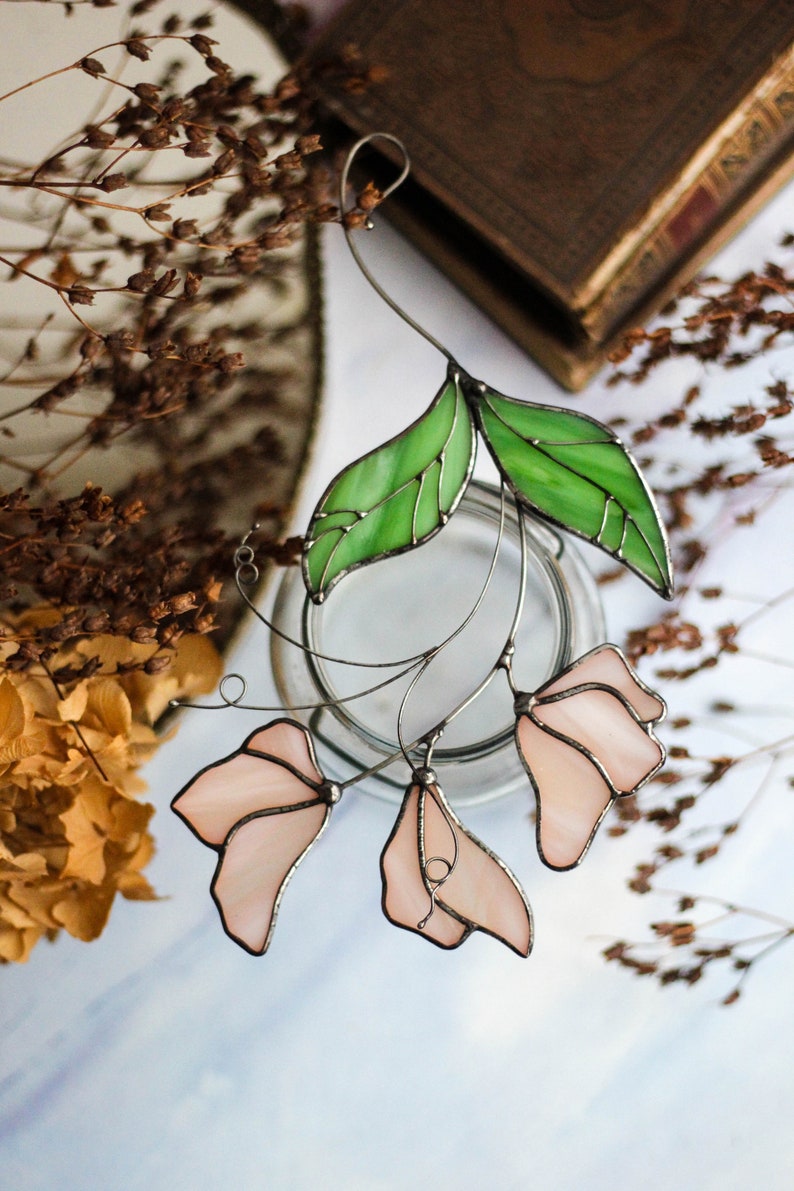 May include: A stained glass floral suncatcher with pink flowers and green leaves. The suncatcher is hanging from a wire and is suspended in front of a white background.