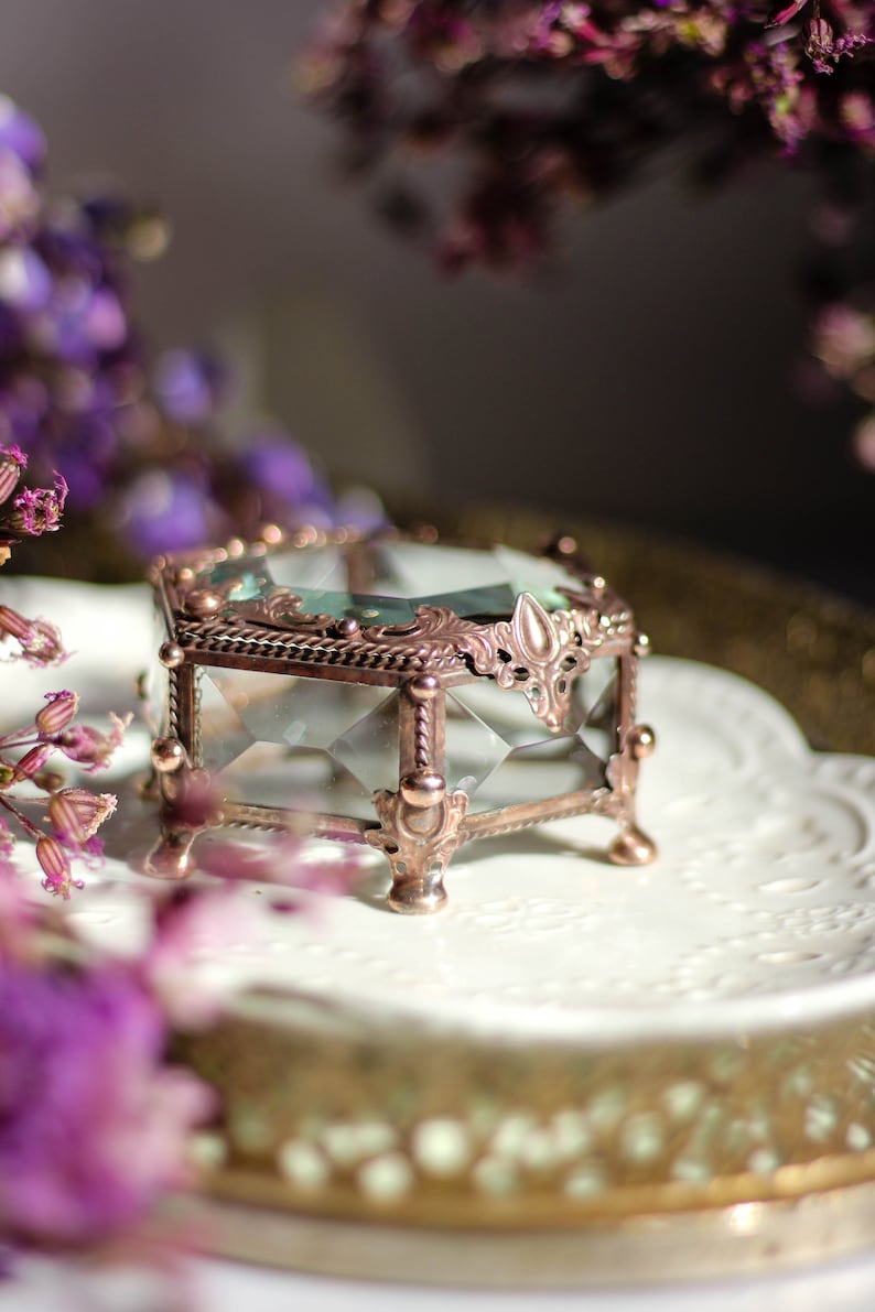 May include: A small, vintage-style glass jewellery box with a copper-coloured metal frame. The box has a faceted glass design and ornate metal detailing. It sits on a white, decorative plate, with purple flowers in the background.