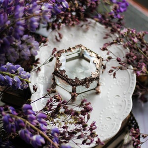 May include: A hexagonal glass and bronze-coloured metal jewellery box with an ornate design rests on a white, patterned plate. The box is surrounded by purple wisteria and dried purple flowers, creating a vintage aesthetic.