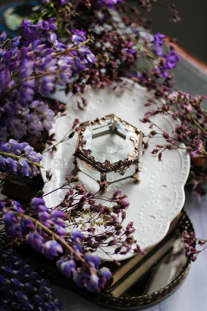 May include: A hexagonal glass and bronze-coloured metal jewellery box with an ornate design rests on a white, patterned plate. The box is surrounded by purple wisteria and dried purple flowers, creating a vintage aesthetic.
