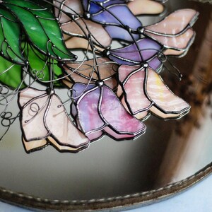 May include: A stained glass window hanging features a cluster of purple, pink, and white flowers with green leaves. The flowers are made of glass and have a delicate, intricate design.