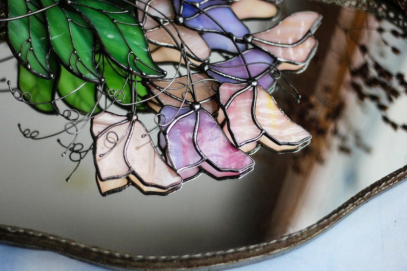 May include: A stained glass window hanging features a cluster of purple, pink, and white flowers with green leaves. The flowers are made of glass and have a delicate, intricate design.
