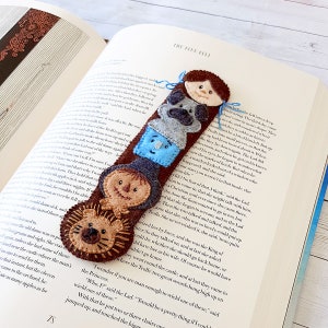 May include: A felt bookmark featuring characters from the book "The Flue Felt". The bookmark is brown with a lion, a scarecrow, a blue character, a grey dog, and a brown-haired person. The bookmark is a fun and whimsical way to mark your place in a book.