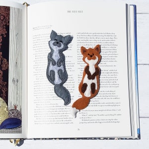 May include: Two felt animal bookmarks: a gray raccoon and a brown fox. The bookmarks are handmade and feature detailed stitching. They are placed on the pages of an open book with the title "The Blue Belt".