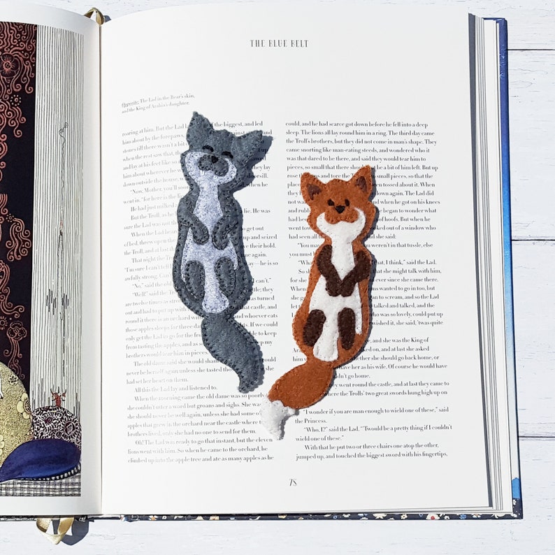 May include: Two felt animal bookmarks: a gray raccoon and a brown fox. The bookmarks are handmade and feature detailed stitching. They are placed on the pages of an open book with the title "The Blue Belt".