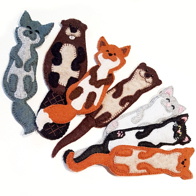 May include: A collection of handmade felt animal bookmarks. The bookmarks include a gray cat, a brown beaver, an orange fox, a brown otter, a gray cat, a black and white cat, and an orange cat. Each bookmark is detailed with stitched features and contrasting felt accents.