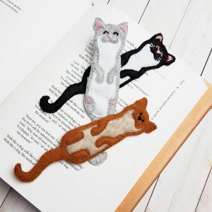 May include: Three handmade felt cat bookmarks in gray, black, and brown with white and tan accents. The bookmarks are lying on an open book. The gray cat has a smiling face. The black cat has a white face and the brown cat has a tan belly.