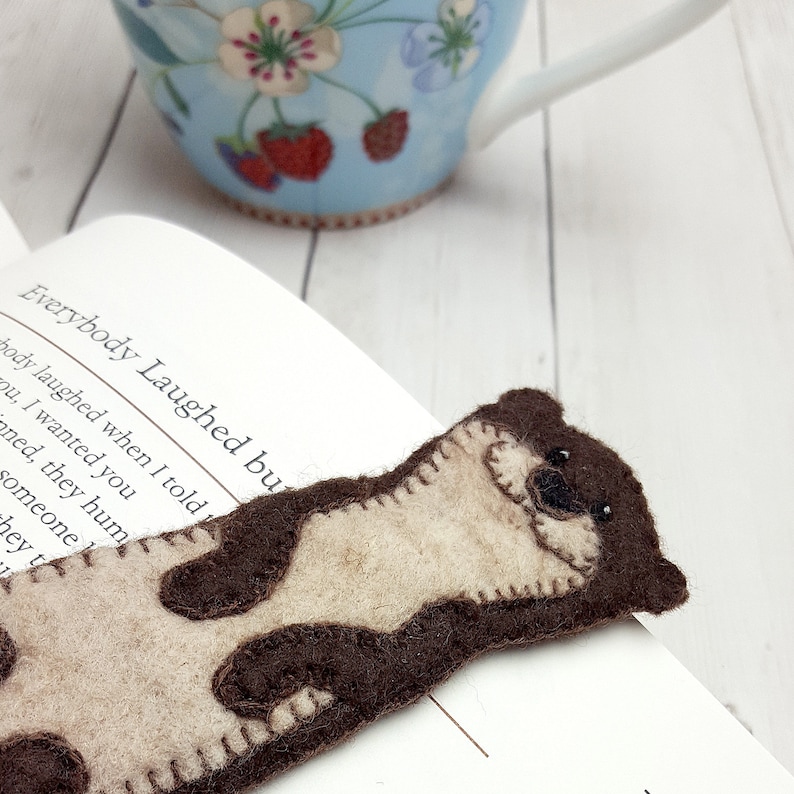 May include: A handmade felt bookmark shaped like an otter, with brown and beige patches. The bookmark is placed on a book with the text "Everybody Laughed bu". A light blue teacup with floral designs is in the background.