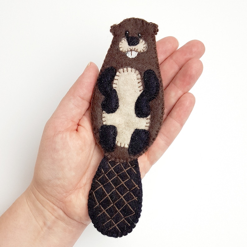 May include: A handmade felt beaver with a brown body, black paws, and a dark brown, crosshatched tail. The beaver has a cream-colored belly and a smiling face with white teeth. The beaver is a whimsical, handcrafted item.